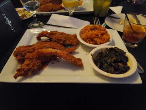 A plate featuring fried chicken strips, rice, and collard greens, served in a dimly lit restaurant setting.