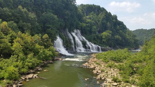 A scenic view of a waterfall cascading into a river, surrounded by lush green trees and rocky terrain.