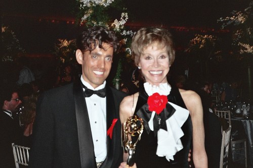 A man in a tuxedo and a woman in a black dress with a red rose pose together, holding an award at a formal event.