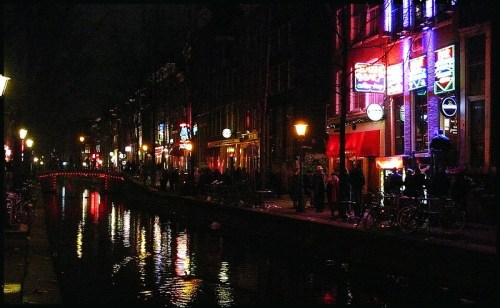 A vibrant canal scene at night, featuring illuminated buildings and reflections in the water, with people and bicycles nearby.