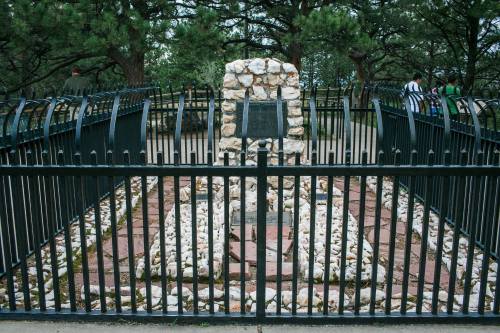 A stone monument surrounded by a black fence, set in a wooded area with visitors in the background.
