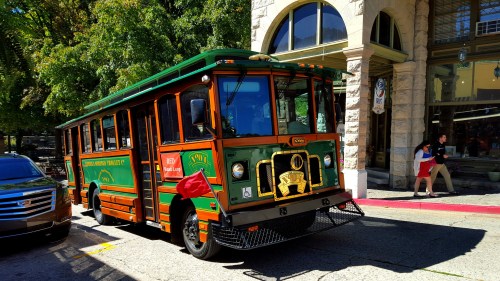 A vintage-style trolley bus parked on a city street, surrounded by trees and buildings.