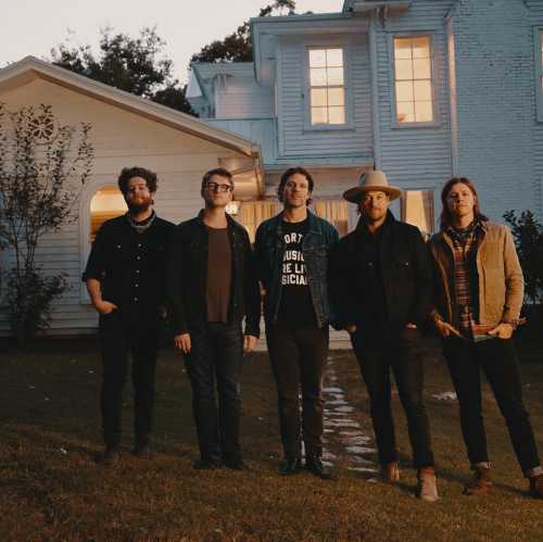 A group of five musicians stands outside a white house at dusk, dressed in casual attire, smiling at the camera.