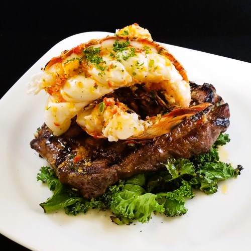 A grilled steak topped with large shrimp, garnished with herbs, served on a bed of kale on a white plate.