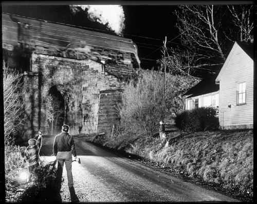 A train passes over a stone bridge at night, with figures holding lanterns on a rural road.
