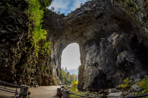 A large rock arch surrounded by greenery, with a pathway leading through the opening and a scenic view beyond.
