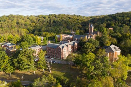 Aerial view of a college campus surrounded by trees and hills, featuring brick buildings and a clock tower.