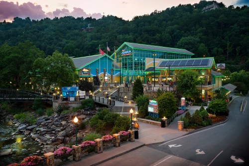A colorful building with large windows, surrounded by greenery and flowers, near a river at sunset.