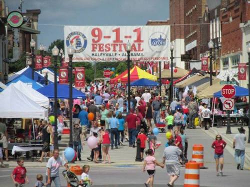 A bustling street festival in Haleyville, Alabama, with colorful tents, people, and a large banner reading "9-1-1 Festival."