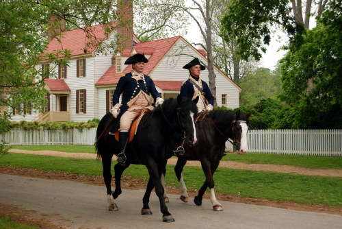 Two men in colonial attire ride horses past a historic house with a white picket fence and greenery.