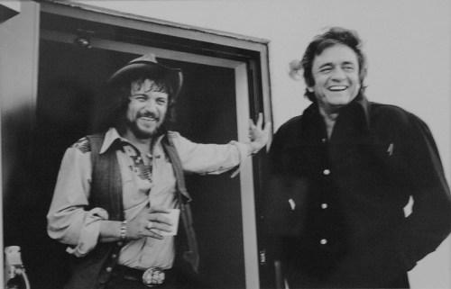 Two smiling men in vintage clothing stand by a doorway, one holding a drink, exuding a relaxed, friendly vibe.