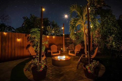 A cozy outdoor seating area with chairs around a fire pit, surrounded by plants and twinkling stars in the night sky.