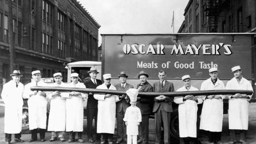 A group of men in white coats and hats holding a giant sausage in front of an Oscar Mayer truck.