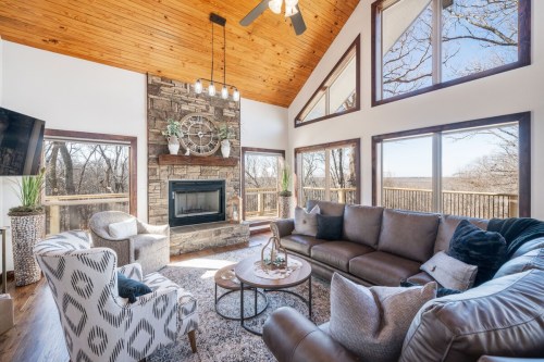 Cozy living room with a stone fireplace, large windows, and a mix of modern and rustic decor. Bright and inviting space.