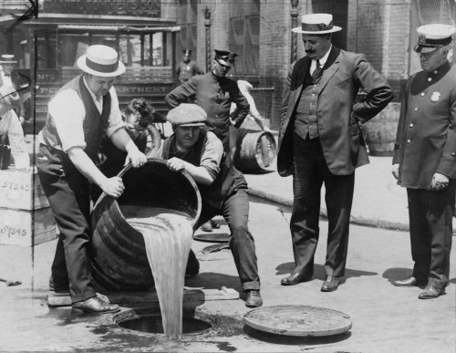 Historical black-and-white photo of men pouring liquid from a barrel into a street, with police officers observing.