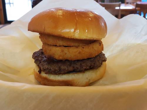 A hamburger with a beef patty, a toasted bun, and a crispy onion ring on top, served on a paper wrapper.