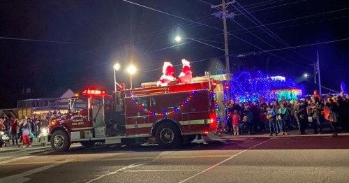 A fire truck decorated with lights, carrying two Santa figures, drives past a crowd during a nighttime holiday parade.