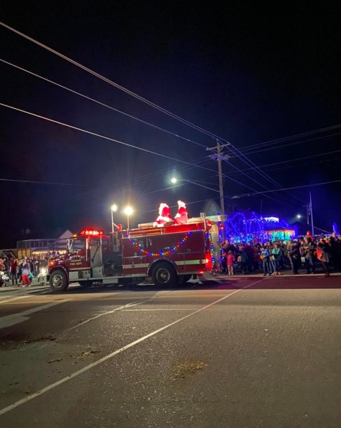 A festive fire truck decorated with lights, carrying two Santa figures, drives past a crowd at night during a holiday parade.