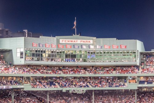 Fenway Park at night, showcasing the iconic stadium facade and flags, with a crowd gathered below.