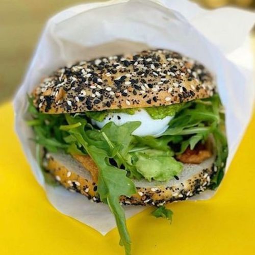A sesame and poppy seed bagel filled with greens, avocado, and a poached egg, wrapped in paper on a yellow surface.