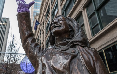 A bronze statue of a woman with raised arm, wearing a coat, set against a city backdrop.