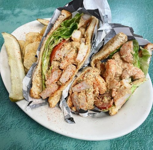 A plate with two shrimp sandwiches on toasted bread, lettuce, and tomato, served with potato chips and pickles.