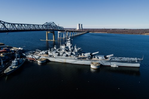 A battleship docked near a bridge and cooling towers, with calm water reflecting the clear sky.