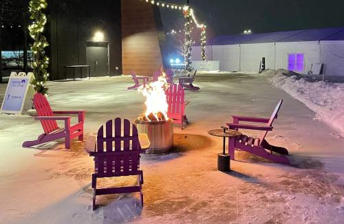A cozy outdoor scene with purple chairs around a fire pit, surrounded by snow and festive lights at night.