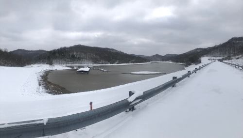 A snowy landscape featuring a lake, a dock, and surrounding hills under a cloudy sky.