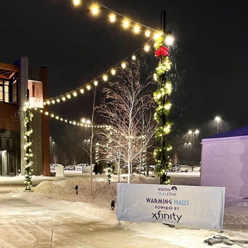 A winter scene with festive lights and a sign for "Warming Haus" powered by Xfinity, surrounded by snow.