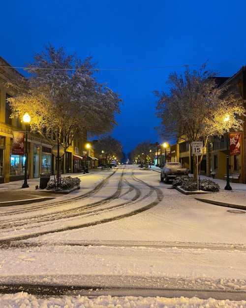Snow-covered street at dusk, lined with trees and buildings, with tire tracks and streetlights illuminating the scene.