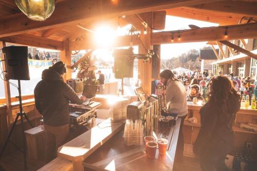 A lively outdoor bar scene with people enjoying drinks, a DJ setup, and sunlight streaming through a wooden structure.