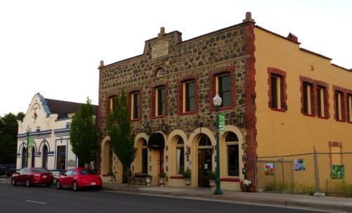 Historic buildings with stone and brick facades, featuring arched windows and greenery, along a quiet street.