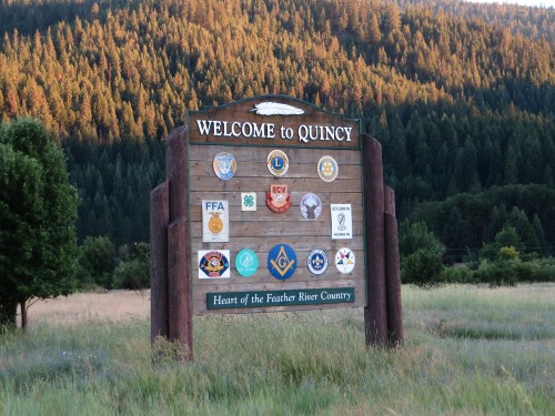 Welcome sign for Quincy, featuring various local organization logos, surrounded by trees and grassy fields.