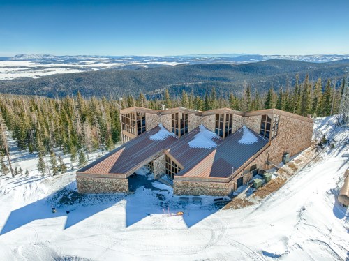 A modern stone building with a metal roof, surrounded by snow-covered mountains and pine trees under a clear blue sky.