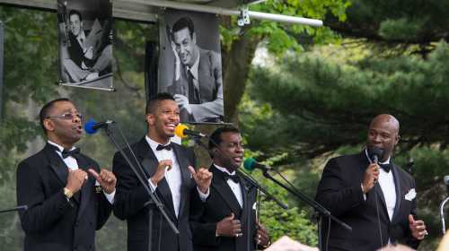 A vocal group in tuxedos performs on stage, with colorful microphones and posters in the background.