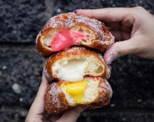 A person holds three stacked, sugar-coated croissants filled with pink, white, and yellow creamy fillings.
