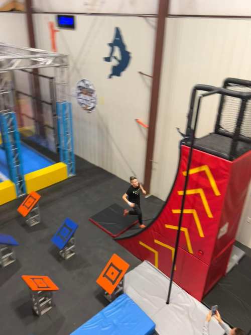 A person runs up a large red ramp in a colorful indoor obstacle course with various platforms and equipment.