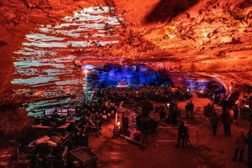 A large crowd gathers in a cave for a concert, with colorful lights illuminating the rock formations above.