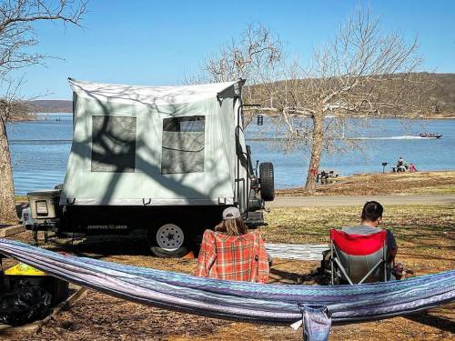 Two people relax in camping chairs by a lake, with a camper and hammock nearby, surrounded by trees and clear blue skies.