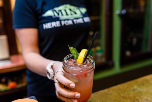 A person holding a mason jar cocktail with a lemon slice and basil, wearing a "Here for the Beer" t-shirt.