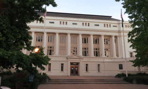A large, classical-style building with columns, flags, and trees in front, illuminated by soft evening light.