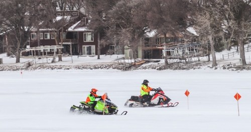 Two snowmobiles ride across a snowy landscape near a lake, with houses visible in the background.