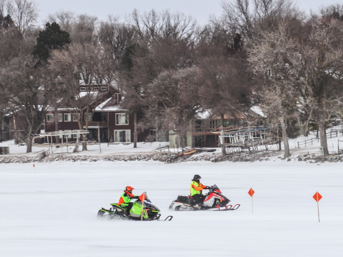 Two snowmobiles race across a snowy landscape near a lake, with houses and trees in the background.