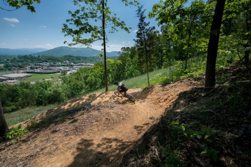 A mountain biker navigates a dirt trail surrounded by trees, with a scenic view of hills and a town in the background.