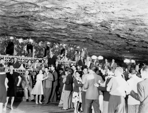 A lively crowd dances and socializes in a cavernous venue with rock walls and decorative lighting.