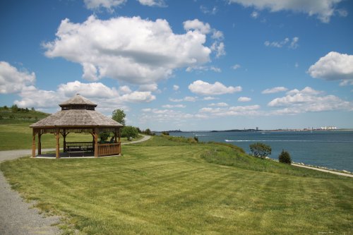 A wooden gazebo on a grassy shore, overlooking a calm body of water under a blue sky with fluffy clouds.