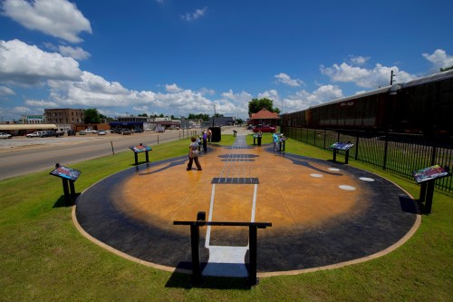 A large guitar-shaped mural on grass, with people walking around and a train in the background under a blue sky.