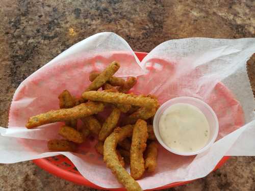 A basket of crispy green bean fries with a small cup of dipping sauce on a textured surface.