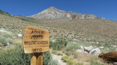 A wooden sign reading "John Muir Wilderness" in front of a mountainous landscape under a clear blue sky.